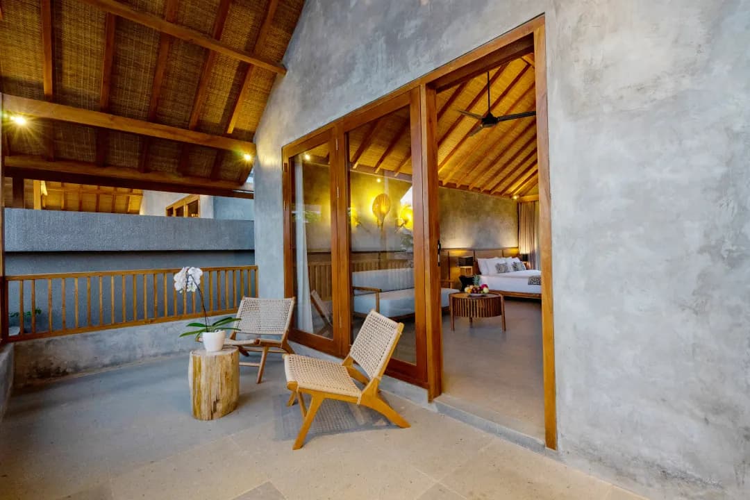 Balcony of bedroom three at Jiwoku Villas Canggu Bali featuring rattan chairs, wooden table, orchid plant, and view into the cozy bedroom interior with warm lighting